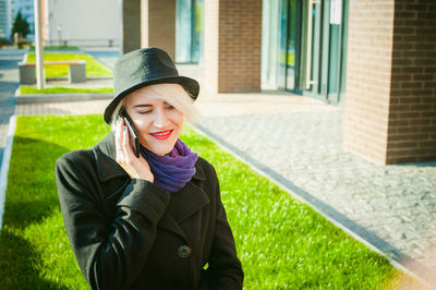 Young woman talking on mobile phone while smoking by built structure