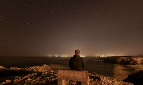 Rear view of man sitting on rock at beach against sky