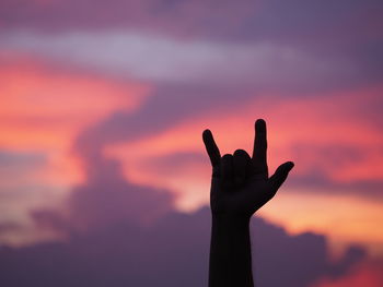 Silhouette cropped hand of man gesturing horn sign against cloudy sky during sunset