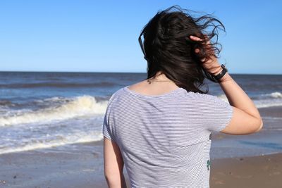 Rear view of woman with tousled hair standing at beach against clear blue sky