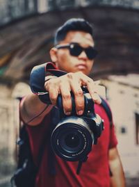 Young man wearing sunglasses photographing with camera while standing against building