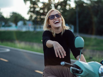 Cheerful young woman wearing sunglasses sitting on motor scooter 
