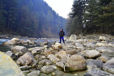 Full length of man standing on rocks at riverbank in forest