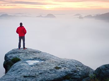 Rear view to traveler stand alone on cliff with mist bellow legs, sunshine in cloudy blue sky