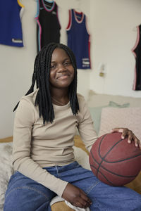 Smiling teenage girl with braided hair holding basketball while sitting in bedroom