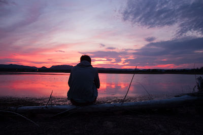 Rear view of silhouette man sitting by lake against sky during sunset