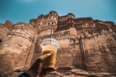 Midsection of person holding drink in a hot summer against a built structure of a fort