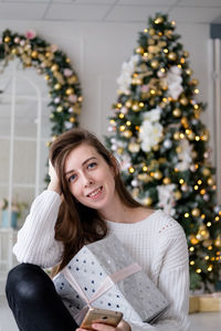 Girl in a white sweater sits with a christmas present on the background of a christmas tree. 