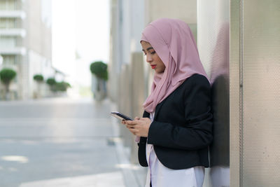 Businesswoman wearing hijab using mobile phone while standing against building