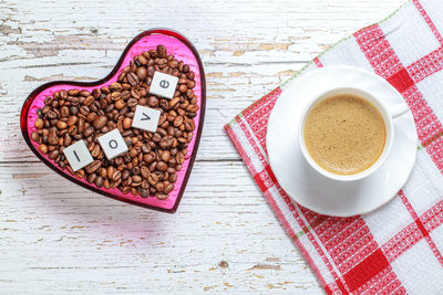 High angle view of cup by heart shaped bowl with roasted coffee beans on table 