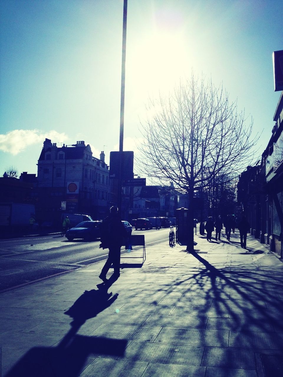 building exterior, architecture, built structure, sunlight, street, transportation, city, car, shadow, men, sun, land vehicle, sky, mode of transport, tree, walking, road, lifestyles, city life