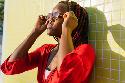 Side view of young african american female in trendy outfit and glasses looking away while standing against yellow background on a sunny day