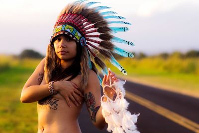 Portrait of shirtless woman in headdress standing against sky