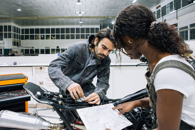 Side view of multiracial male and female mechanics repairing custom motorbike in spacious workshop while working together