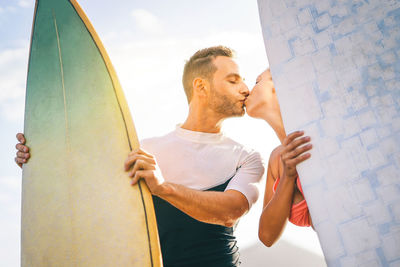 Man and woman kissing and holding surfboards while standing against sky