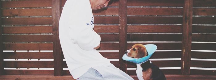 Midsection of man with dog wearing protective collar by wooden wall