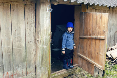 Village boy in boots stands at the door of the barn in warm clothes