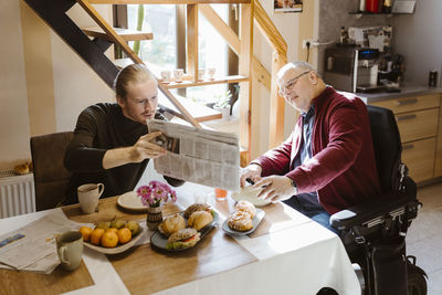 Young man showing newspaper to senior father with disability while eating breakfast at home