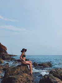 Side view of young woman sitting on rock by sea against sky