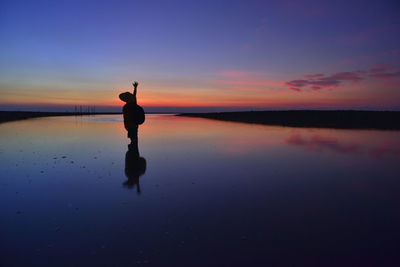 Silhouette man standing in shallow water at beach against sky during sunset