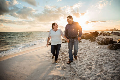 Rear view of couple standing at beach against sky during sunset