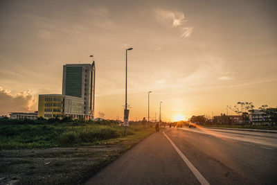 Road by buildings against sky during sunset