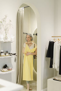 Smiling woman holding yellow dress stepping out from dressing room at store