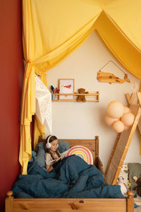 Cute concentrated little girl with long hair in pajama and headphones using stylus while drawing on graphic tablet and sitting on comfortable bed under warm blanket at home