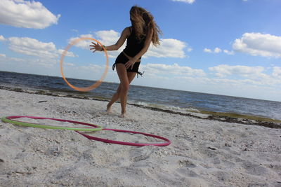 Full length of young woman playing with plastic hoop at beach against cloudy sky