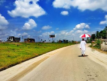 Rear view of man holding umbrella while walking on road against cloudy sky