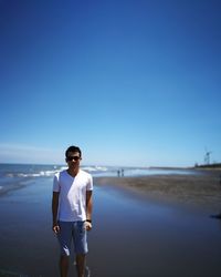 Portrait of young man wearing sunglasses standing at beach against blue sky during sunny day