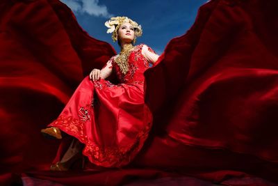 Low angle view of beautiful model wearing red period costume while sitting amidst curtain