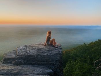 Portrait of smiling woman sitting on cliff by sea against sky during sunset