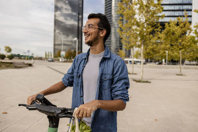 Happy young man with electric push scooter standing at footpath