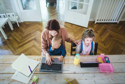 High angle view of mother working on laptop while sitting with cute son by daughter using digital tablet at table