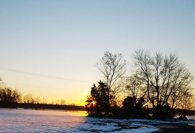 Silhouette trees on frozen landscape against clear sky during sunset