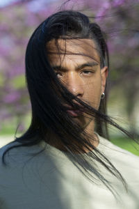 Handsome young hispanic male with long black hair looking at camera while standing in park with blooming trees on blurred background