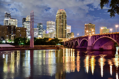 Illuminated bridge over river by buildings against sky at night