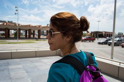 Young woman carrying backpack on street in city against sky