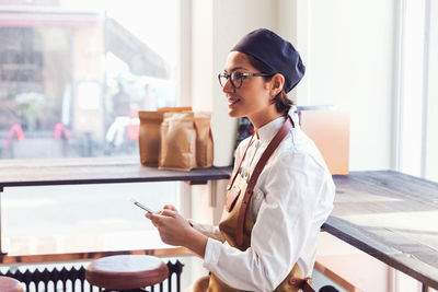 Smiling female owner holding smart phone while standing by window in grocery store