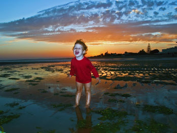Full length of boy shouting at beach against cloudy sky during sunset