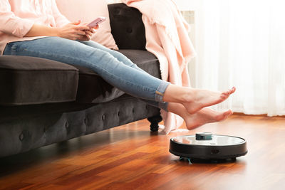 Low section of woman sitting on sofa with robotic vacuum cleaner at home