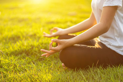 Low section of woman meditating while sitting on field at park during sunset