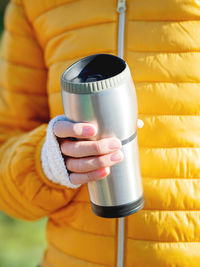 Women in bright yellow jacket is holding thermos mug. hot tea or other beverage on cool autumn day.