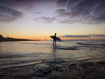 Silhouette mature man with surfboard standing at beach against sky during sunset