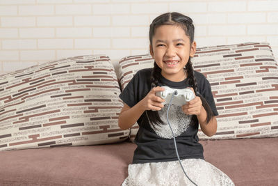 Portrait of smiling girl holding camera while sitting against wall