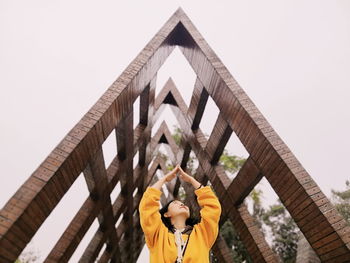 Low angle view of young woman with arms raised standing against built structure