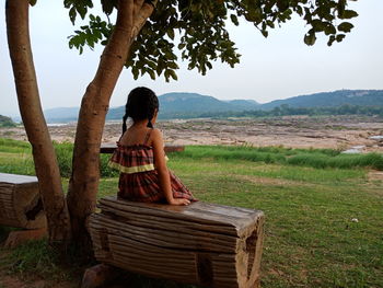 Girl sitting on seat by tree against sky