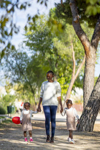 Full body of young stylish african american mother holding hands of cute little twin girls while walking together in park on sunny day