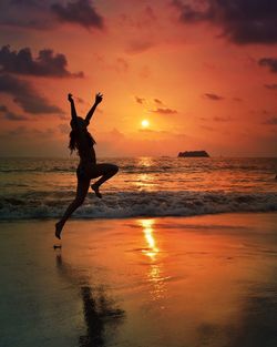 Silhouette woman with arms raised jumping at beach against sky during sunset